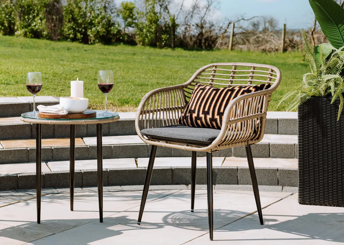 A bistro set with 2 wicker chairs and a small glass table from EZ Livings Menorca range. Angled view of rattan chair with metal legs
