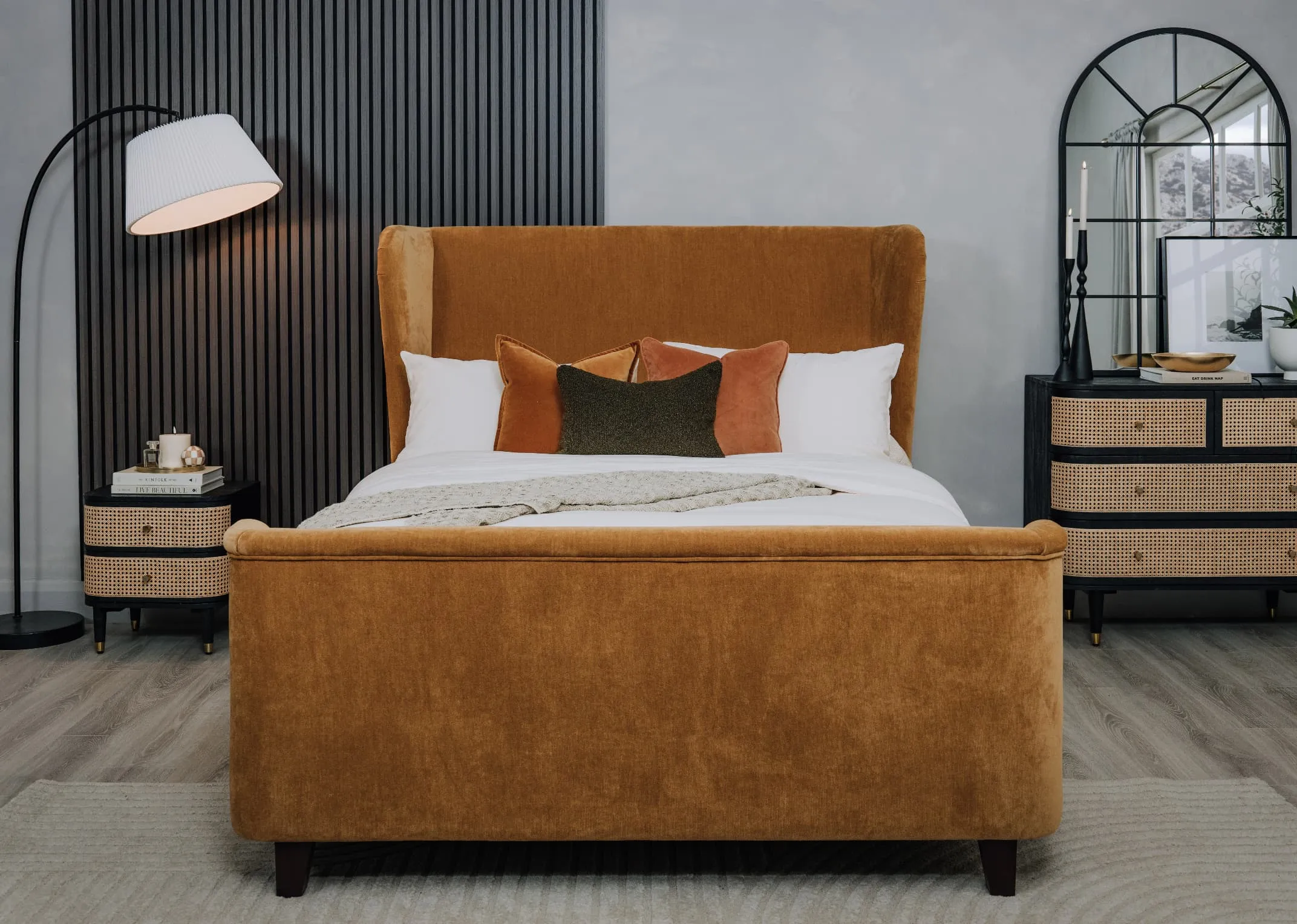 A lifestyle image of the Maison gold bedframe with Black panel wall and lenny locker and chest with the Belle mirror and Arc lamp.